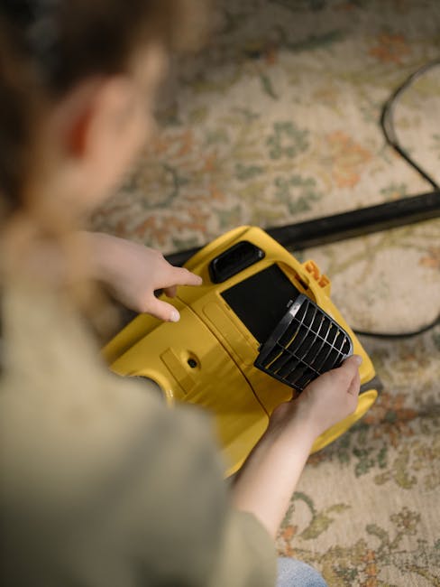 A person dressed in a light-colored jumpsuit is operating a yellow vacuum cleaner with a long hose in a dark room. The individual is bent forward, guiding the cleaning equipment across a carpeted surf
