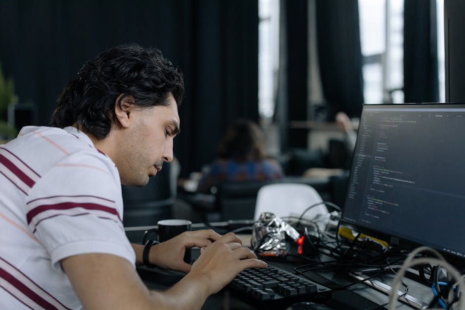 A young man with dark, curly hair and light skin is seated at a desk in an office environment, focused on a desktop computer with a large monitor displaying lines of code or programming text. The desk