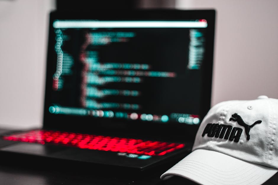A young man with dark, curly hair and light skin is seated at a desk in an office environment, focused on a desktop computer with a large monitor displaying lines of code or programming text. The desk