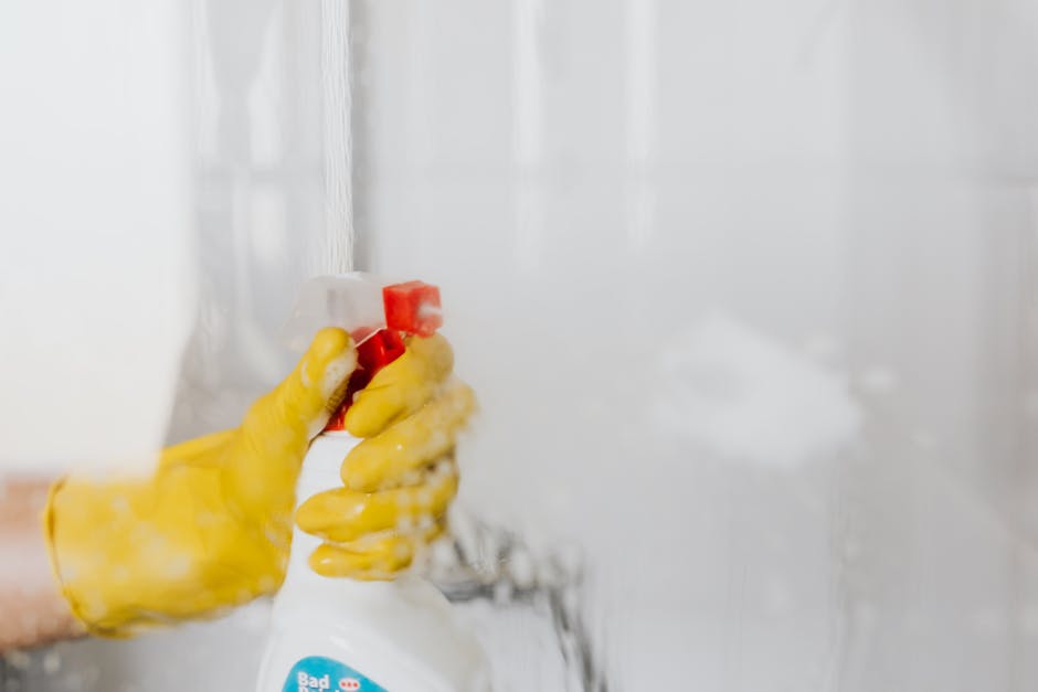 Three professional cleaners from Cleaners Hackney stand indoors in a modern room with neutral-colored walls, wearing matching grey t-shirts and bright orange work overalls. They are holding cleaning t